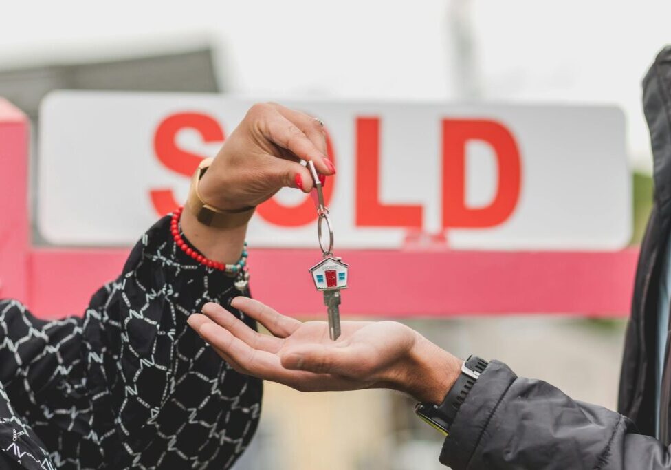 Hands exchanging house keys with a 'SOLD' sign in the background.